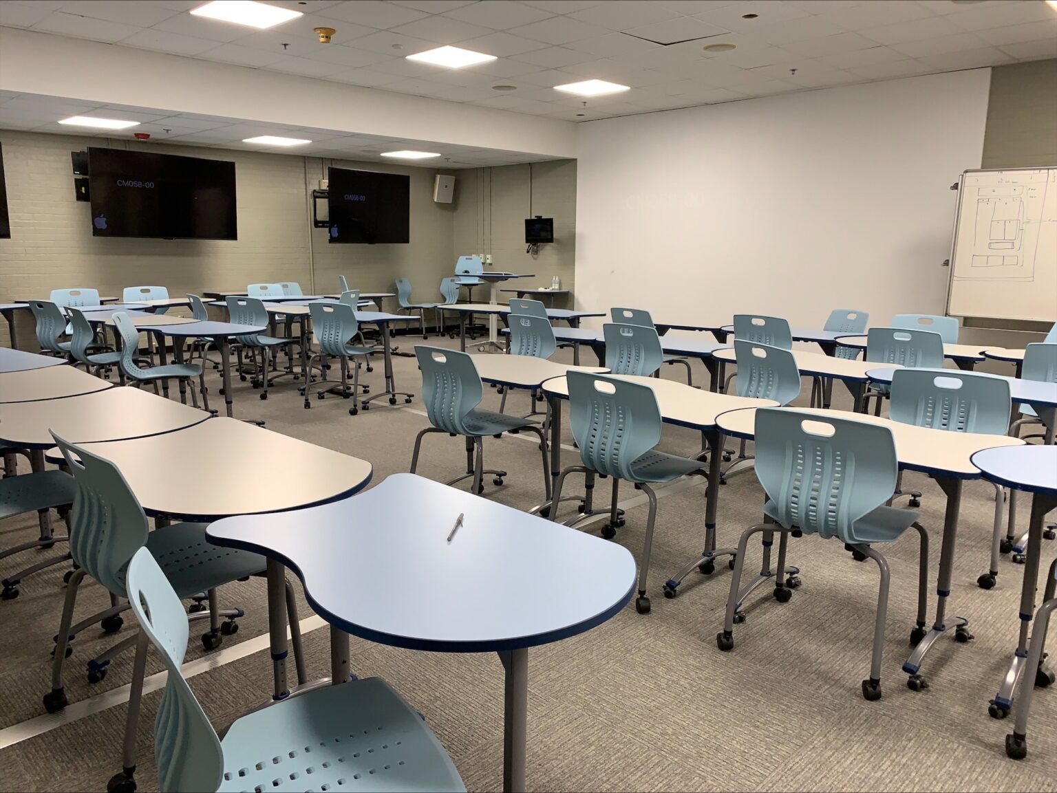 Interior of a classroom facing a large board, with mounted TVs and lots of modular desks. 