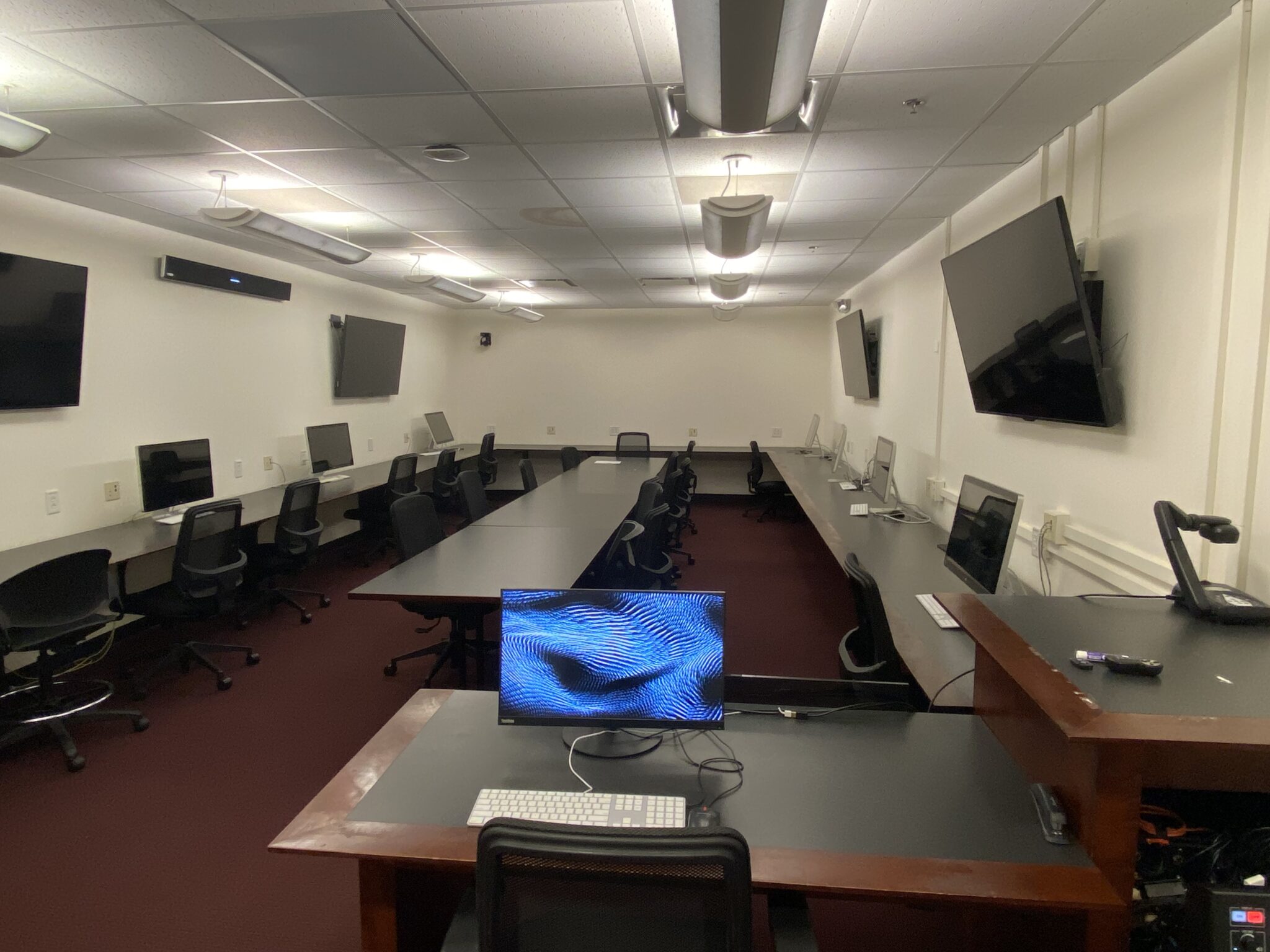 Classroom interior with desks and monitors in a ring and a conference table in the middle.