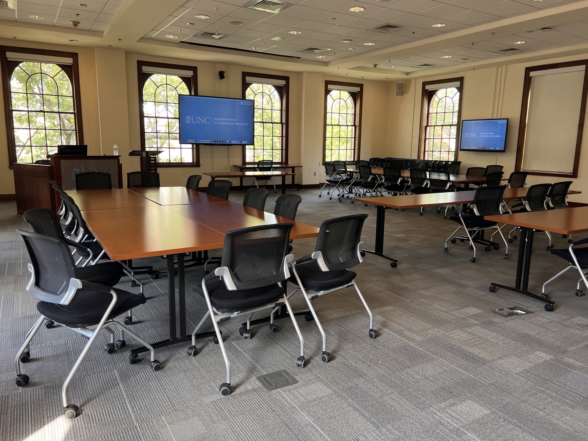 Conference room with mounted monitors and several groupings of tables surrounded by rolling chairs.